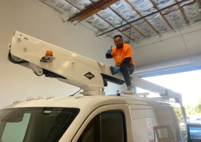 Worker in orange shirt giving thumbs up while installing Versalift equipment on white utility truck.