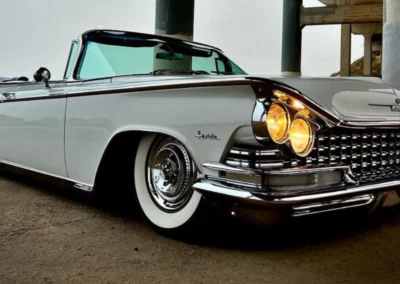 A vintage white Buick convertible with glowing headlights parked under a bridge on a cloudy day.